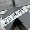 A '2D PRINT' number plate with standard printed black characters on a white plate, resting on a dark, circular table.