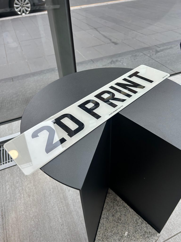 Close-up of a 2D printed UK number plate displaying '2D PRINT' resting on a modern black table.