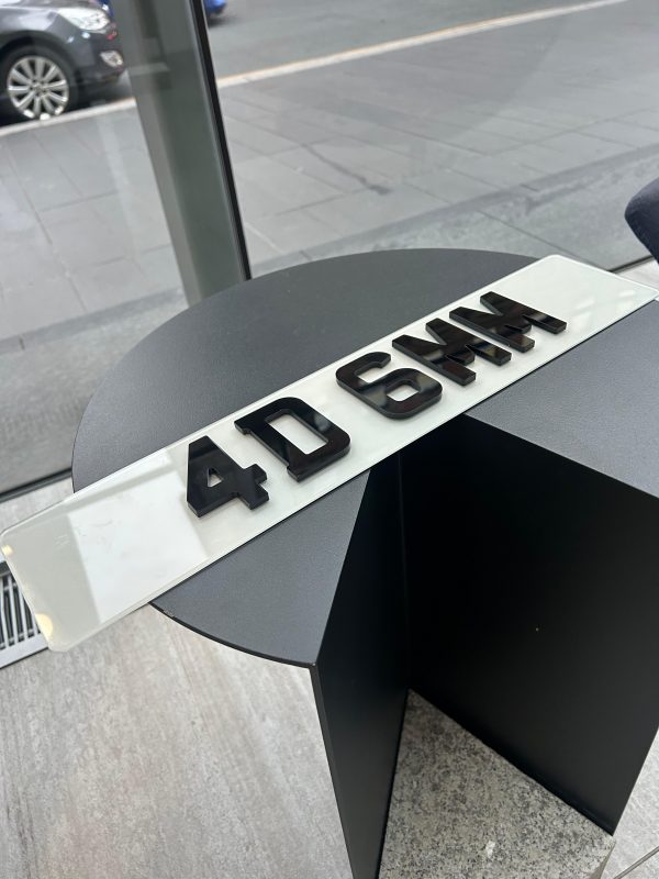 A 4D 6mm number plate with raised black acrylic characters on a white plate, resting on a dark, circular table.
