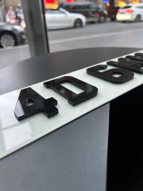 An angled close-up of a '4D GEL' number plate, showcasing the raised, glossy black characters against a blurred street background.